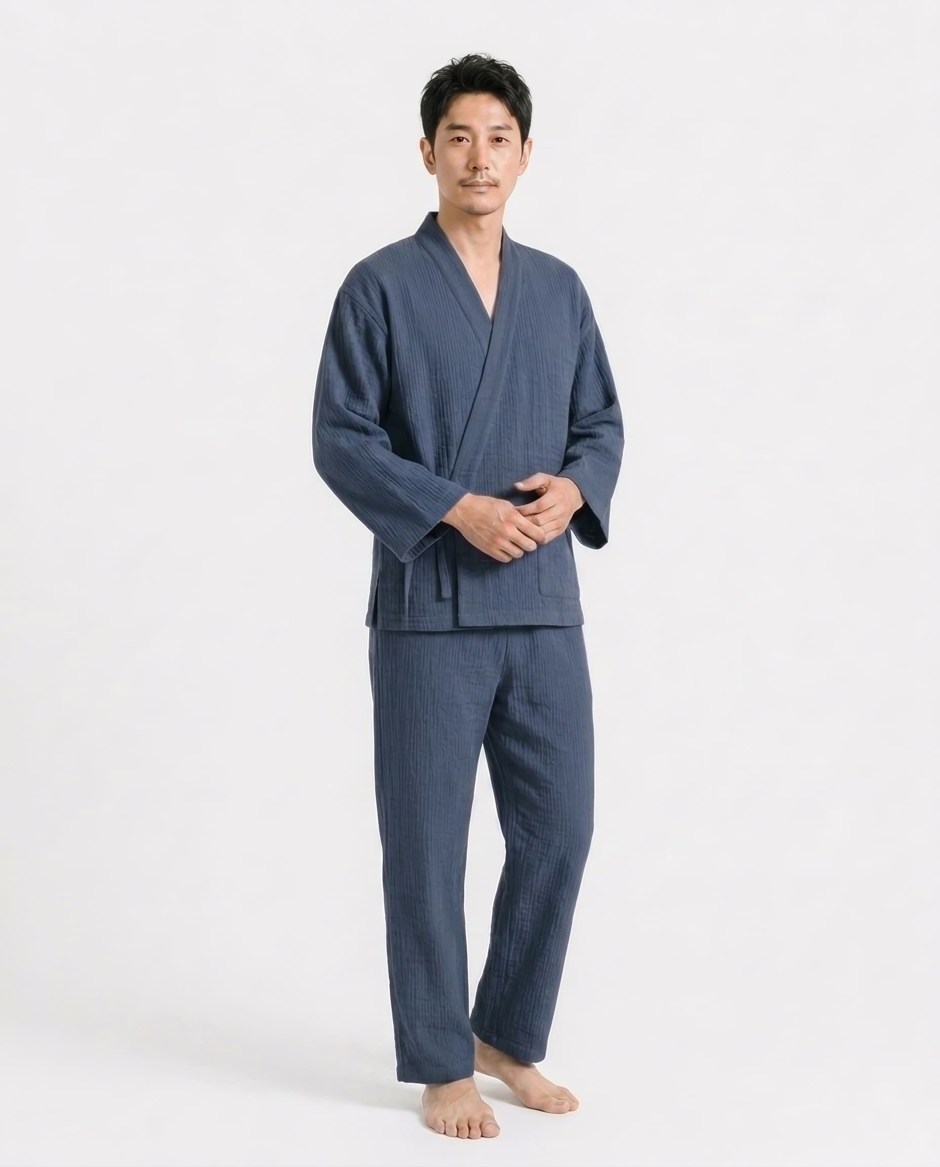 A man wearing a dark blue two-piece robe set consisting of a long-sleeve wrap jacket and matching pants, standing against a plain white studio background.