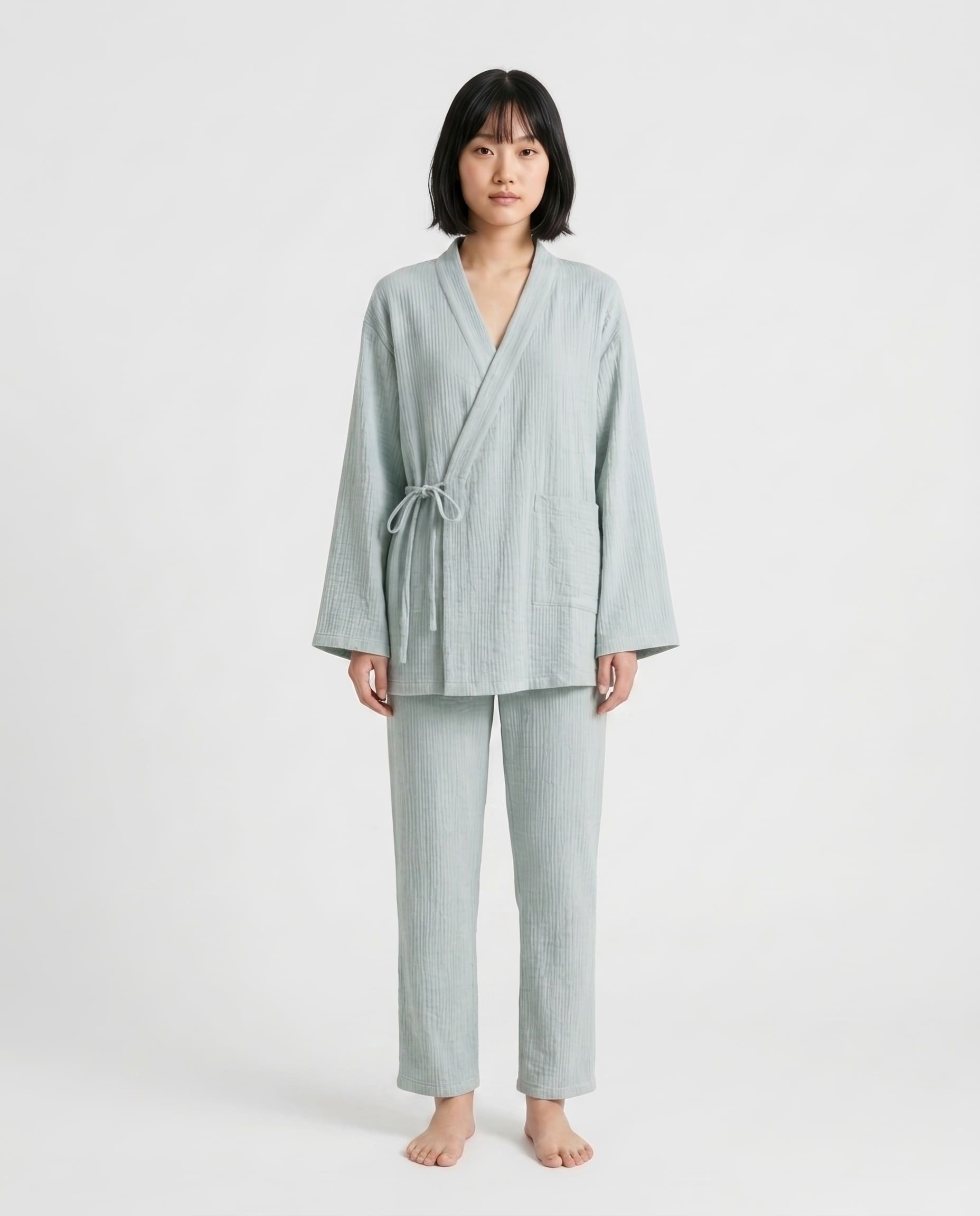 A woman stands barefoot against a plain light gray studio background wearing a matching light sage green pajama set consisting of a long-sleeve wrap robe with a tie waist and coordinating straight-leg pants.