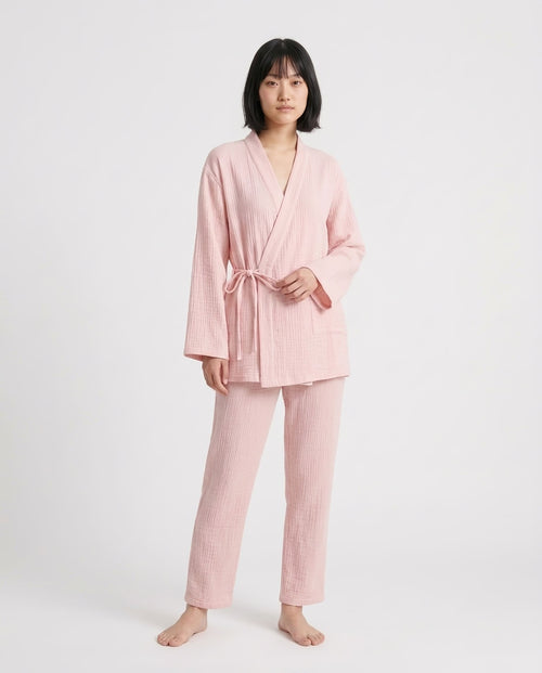A woman modeling a light pink two-piece pajama set with a textured waffle fabric, consisting of a long-sleeve tie-front robe and matching pants, against a plain light gray studio background.